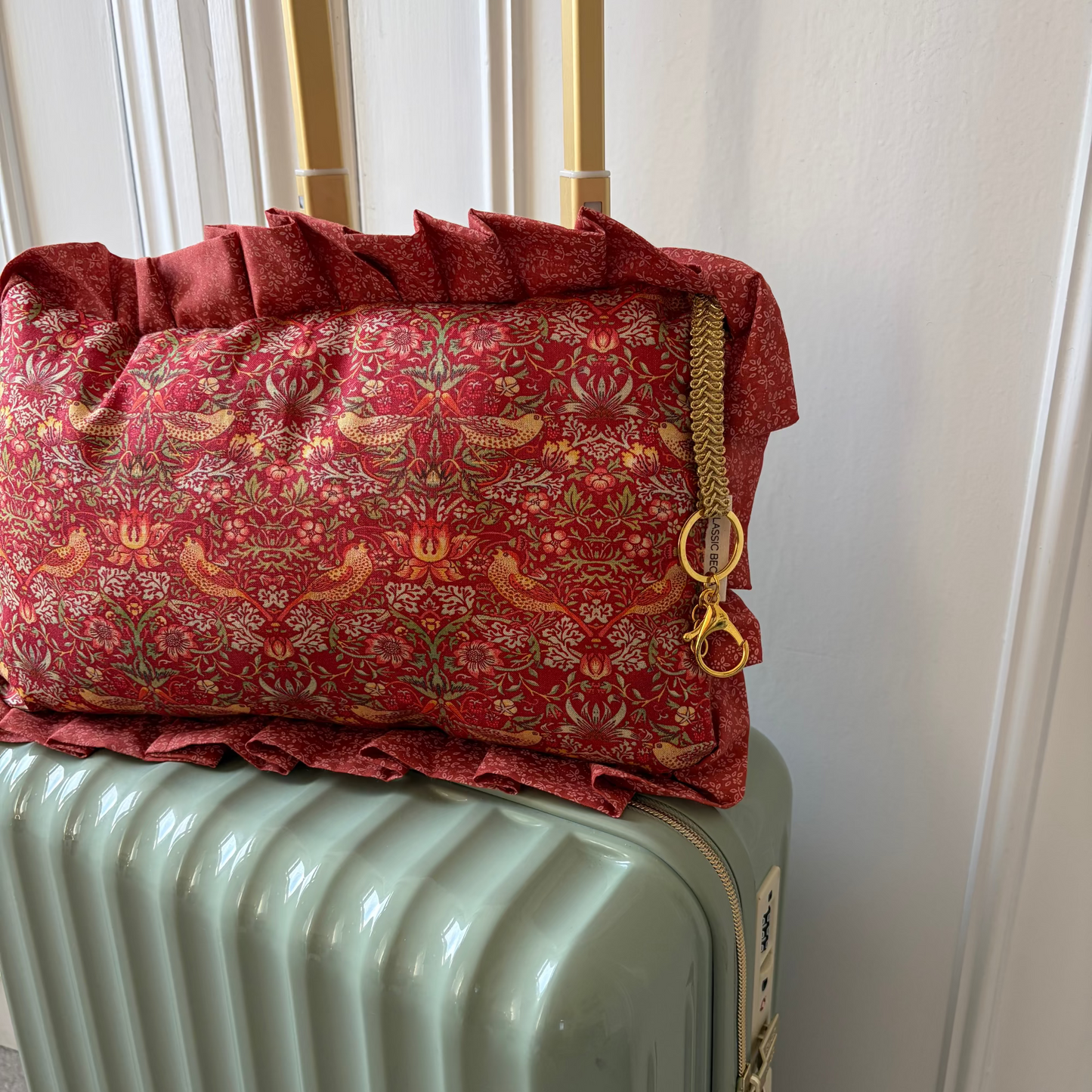 Red william morris Travel Bag with gold accents on a green suitcase against a white door background